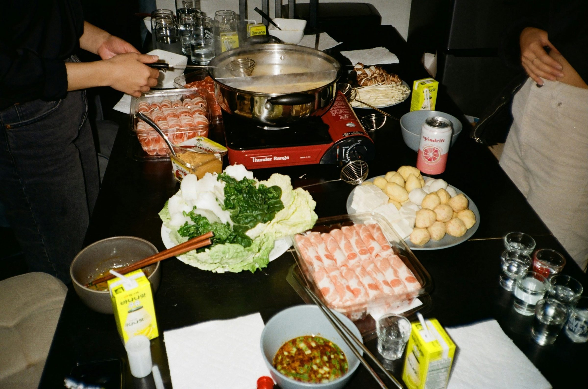 A hotpot dinner: a steaming pot on a table surrounded by plates of meat, fish balls, tofu and vegetables